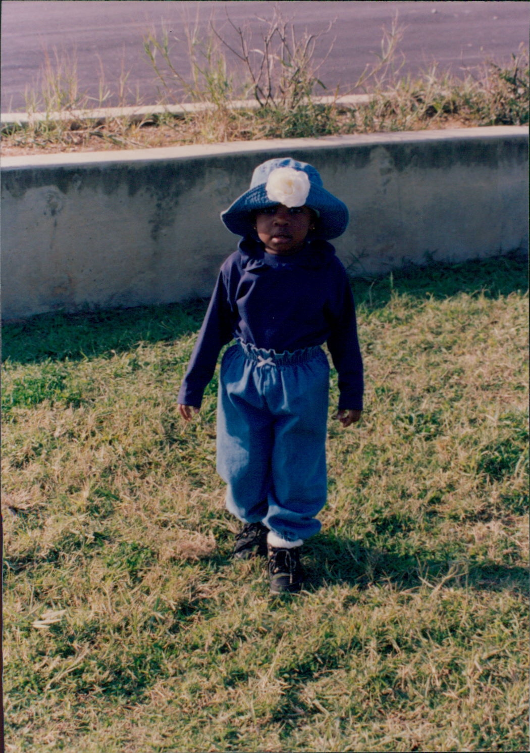 A young girl in a denim bucket hat and denim overalls walking on grass